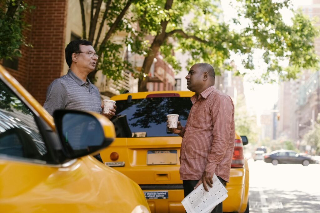 Side view of multiracial male coworkers with newspaper and takeaway coffee speaking near taxi autos while looking at each other in town